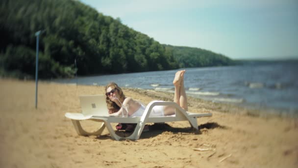 Una joven en un bikini blanco se encuentra y broncea en una tumbona en una playa de arena de mar y está trabajando en un ordenador portátil — Vídeo de stock