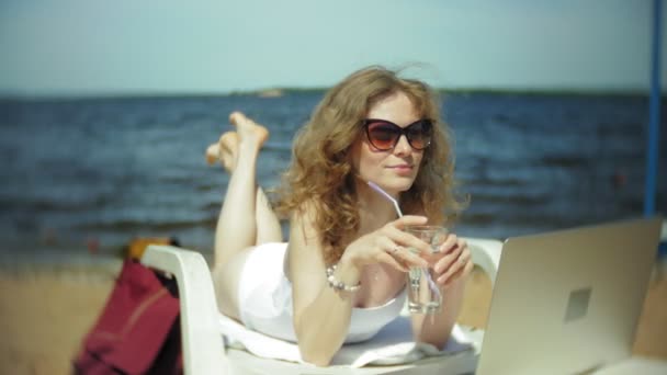 Een jong meisje in een witte bikini ligt en tans op een strandstoel op een zee zand strand en is bezig met een laptop — Stockvideo