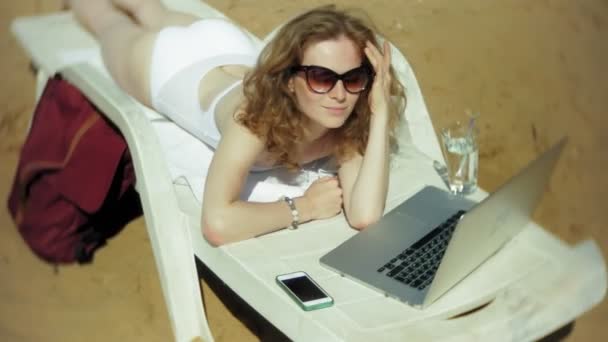 Молода дівчина в білому бікіні лежить і пил на шезлонгу на морському піщаному пляжі і працює на ноутбуці — стокове відео