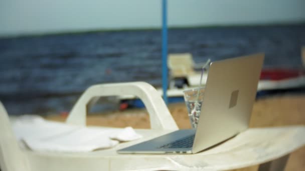 Bärbar dator och ett glas vatten på en schäslong med utsikt över havet — Stockvideo
