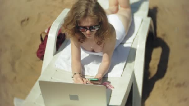 Una joven en un bikini blanco se encuentra y broncea en una tumbona en una playa de arena de mar y está trabajando en un ordenador portátil — Vídeo de stock