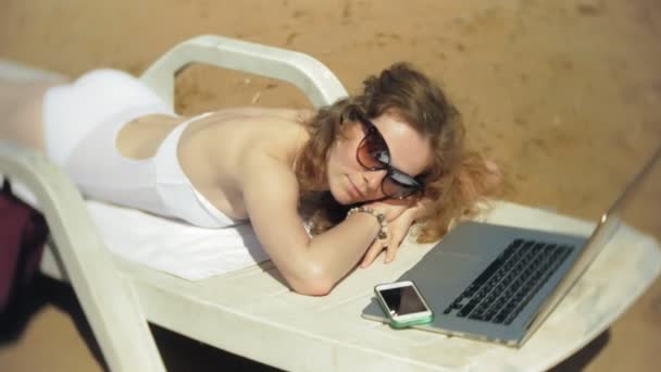 Ένα νεαρό κορίτσι σε ένα λευκό μπικίνι βρίσκεται και μαυρίσματα σε μια ξαπλώστρα στην αμμώδη παραλία και εργάζονται σε ένα φορητό υπολογιστή — Αρχείο Βίντεο