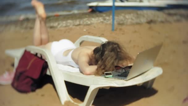 Egy fiatal lány, egy fehér bikiniben fekszik és varádics a nyugágyon a tenger homokos strandján és működő-ra egy laptop — Stock videók