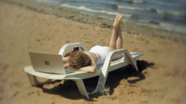 Egy fiatal lány, egy fehér bikiniben fekszik és varádics a nyugágyon a tenger homokos strandján és működő-ra egy laptop — Stock videók