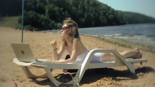 흰색 비키니에 젊은 여자와 바다 모래 해변에 해먹이에 담배 있으며 랩톱에서 작동 — 비디오