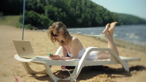 Una joven en un bikini blanco se encuentra y broncea en una tumbona en una playa de arena de mar y está trabajando en un ordenador portátil — Vídeos de Stock