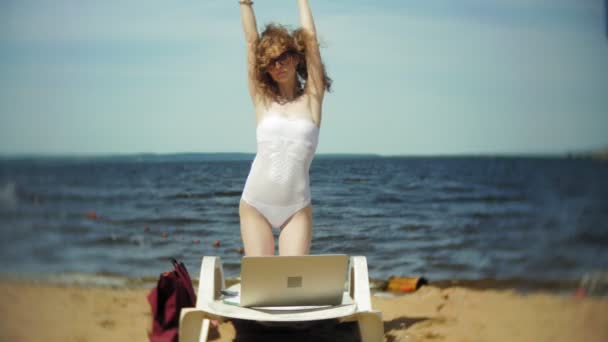 Beyaz bikinili genç bir kız yatıyor ve tans üzerinde bir deniz kumlu plajda şezlong ve bir dizüstü bilgisayar üzerinde çalışan — Stok video