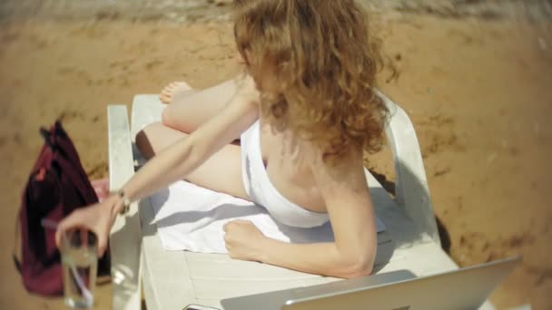 Une jeune fille en bikini blanc se couche et prend un bain de soleil sur une chaise longue sur la plage de sable de la mer et boit un cocktail — Video