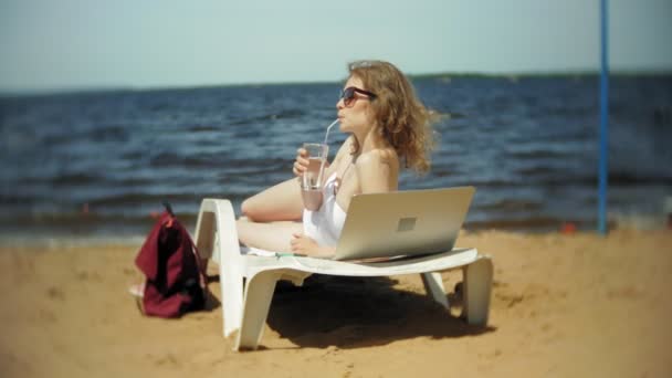 Ein junges Mädchen im weißen Bikini liegt und sonnt sich auf einer Liege am Sandstrand des Meeres und trinkt einen Cocktail — Stockvideo