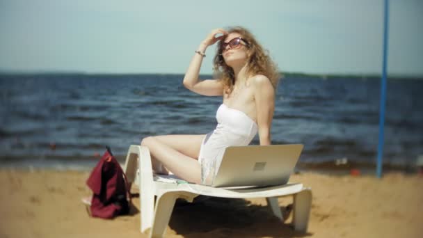 白ビキニの若い女の子がある海砂浜ビーチでデッキチェアに日焼けし、はラップトップに取り組んで — ストック動画