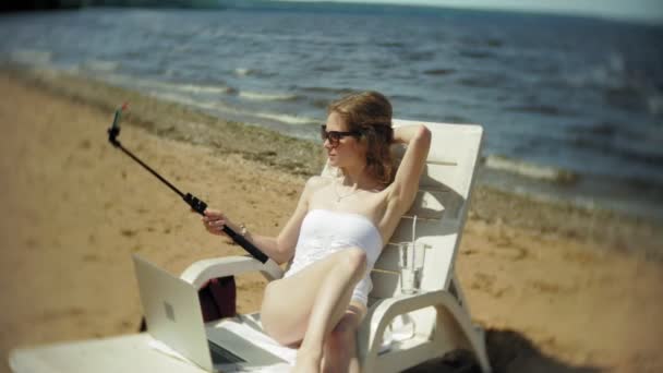 Una joven en bikini blanco se encuentra y toma el sol en una tumbona en una playa de arena de mar y fotografías en un teléfono inteligente SELFI — Vídeos de Stock