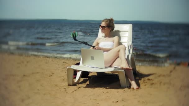 Una joven en bikini blanco se encuentra y toma el sol en una tumbona en una playa de arena de mar y fotografías en un teléfono inteligente SELFI — Vídeos de Stock