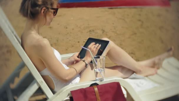 白ビキニの若い女の子がある海砂浜ビーチでデッキチェアに日焼けし、は、タブレットに取り組んで — ストック動画
