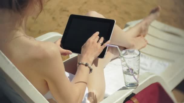 Egy fiatal lány, egy fehér bikiniben fekszik és varádics a nyugágyon a tenger homokos strandján és működő-ra egy tabletta — Stock videók