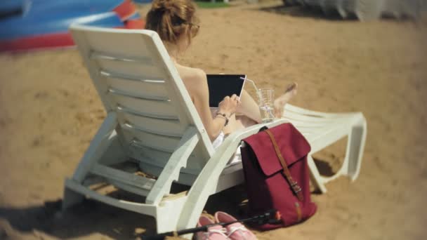 Una joven en un bikini blanco se encuentra y broncea en una tumbona en una playa de arena de mar y está trabajando en una tableta — Vídeo de stock