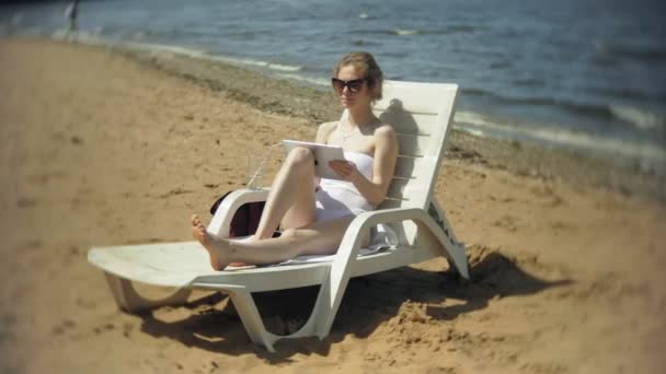 Une jeune fille en bikini blanc se couche et bronzait sur un transat sur une plage de sable de mer et travaille sur une tablette — Video