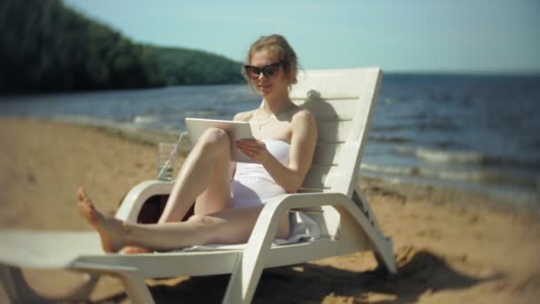 Ein junges Mädchen im weißen Bikini liegt und bräunt auf einem Liegestuhl am Sandstrand und arbeitet an einem Tablet — Stockvideo