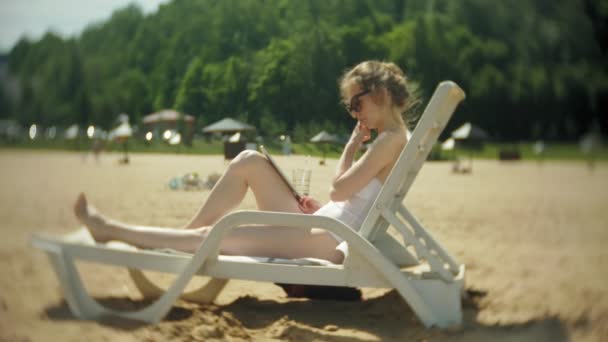 Ein junges Mädchen im weißen Bikini liegt und bräunt auf einem Liegestuhl am Sandstrand und arbeitet an einem Tablet — Stockvideo