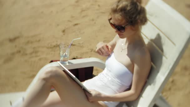 Молода дівчина в білому бікіні лежить і пил на шезлонгу на морському піщаному пляжі і працює на планшеті — стокове відео