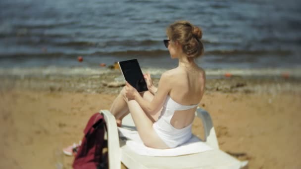 Egy fiatal lány, egy fehér bikiniben fekszik és varádics a nyugágyon a tenger homokos strandján és működő-ra egy tabletta — Stock videók