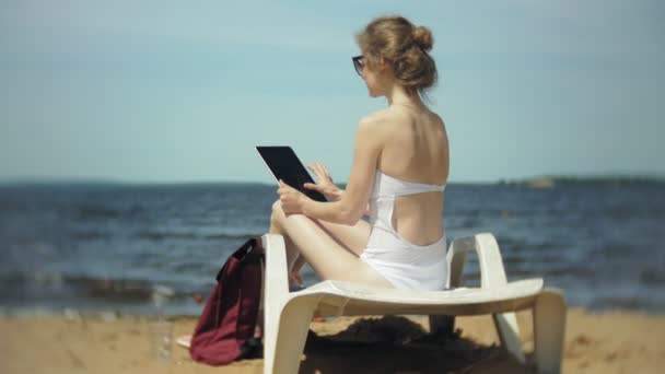 Una joven en un bikini blanco se encuentra y broncea en una tumbona en una playa de arena de mar y está trabajando en una tableta — Vídeo de stock