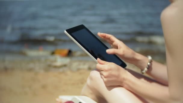 Una joven en un bikini blanco se encuentra y broncea en una tumbona en una playa de arena de mar y está trabajando en una tableta — Vídeos de Stock