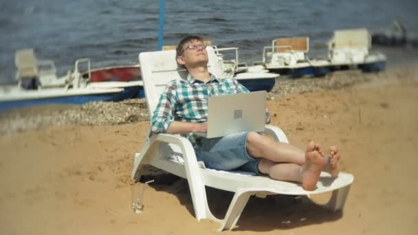 Een jong meisje in een witte bikini ligt en tans op een strandstoel op een zee zand strand en is bezig met een laptop — Stockvideo