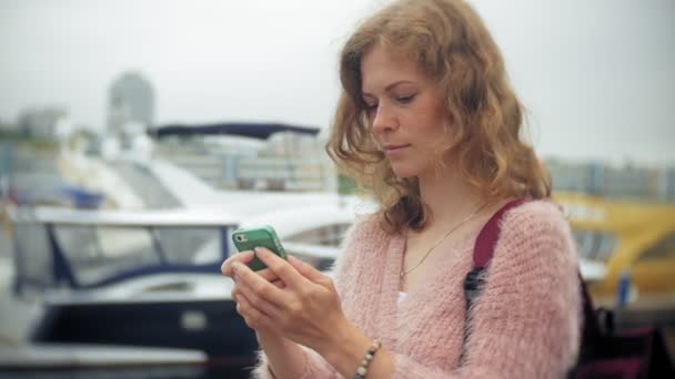 Uma menina usando um smartphone à beira-mar, iate e vela no porto . — Vídeo de Stock