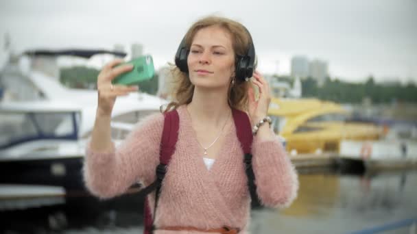 女の子がビーチでスマート フォンを使用してヘッドフォンで音楽を聴く、ダンス、ヨット、港でのセーリング. — ストック動画