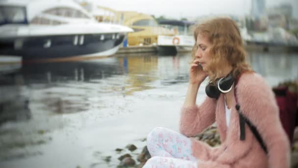 一个女孩使用智能手机在海滩上, 听音乐, 在耳机, 跳舞的游艇和帆船在海港. — 图库视频影像
