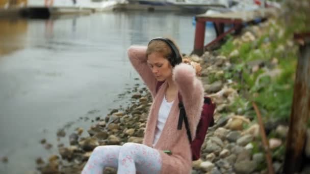 Uma menina usando um smartphone na praia, ouvindo música em fones de ouvido, dançando um iate e navegando no porto . — Vídeo de Stock