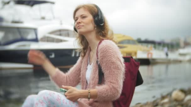 Ένα κορίτσι με ένα smartphone στην παραλία, να ακούτε μουσική στα ακουστικά, χορεύουν ένα σκάφος και ιστιοπλοΐα στο λιμάνι. — Αρχείο Βίντεο
