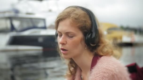 女の子がビーチでスマート フォンを使用してヘッドフォンで音楽を聴く、ダンス、ヨット、港でのセーリング. — ストック動画