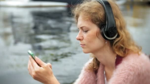 Дівчина, використовуючи смартфон на пляжі, слухає музику в навушниках, танцює яхту і пливе в гавані . — стокове відео