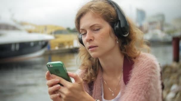 Een meisje met een smartphone op het strand, luisteren naar muziek in hoofdtelefoons, dansen van een jacht en zeilen in de haven. — Stockvideo