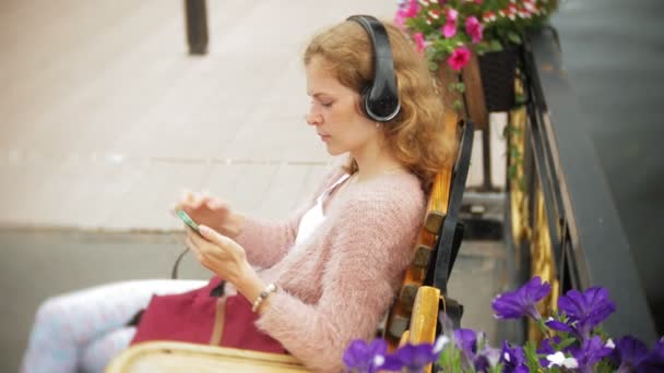 Kız çıtır sahilde, bir bankta bir smartphone kullanarak kulaklık müzik dinlemek, bir yat dans ve limanda yelken. — Stok video