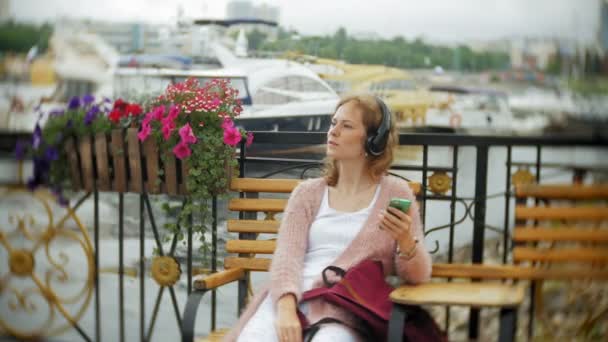 Девушка с помощью смартфона на скамейке в цветах на пляже, слушает музыку в наушниках, танцует яхту и плавает в гавани . — стоковое видео