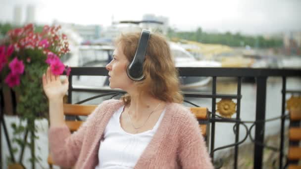 Een meisje met een smartphone op een bankje in de bloemen op het strand, luisteren naar muziek in hoofdtelefoons, dansen van een jacht en zeilen in de haven. — Stockvideo