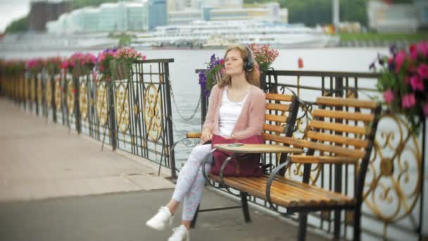 Una chica usando un smartphone en un banco con flores en la playa, escuchando música en auriculares, bailando un yate y navegando en el puerto . — Vídeo de stock