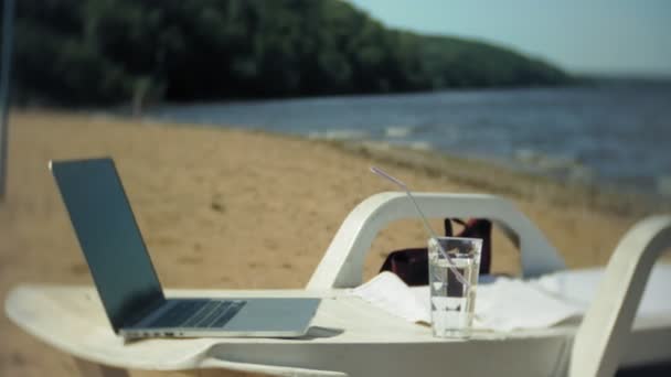 Bärbar dator och ett glas vatten på en schäslong med utsikt över havet — Stockvideo