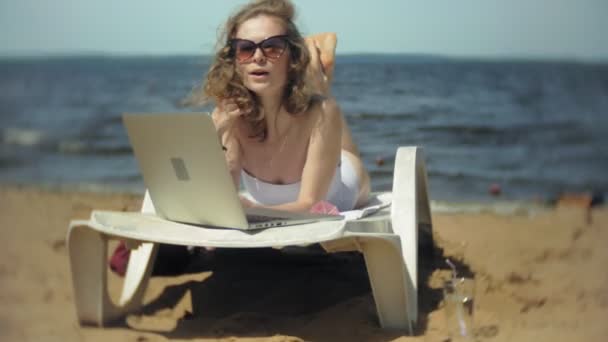 Una joven en un bikini blanco se encuentra y broncea en una tumbona en una playa de arena de mar y está trabajando en un ordenador portátil — Vídeo de stock