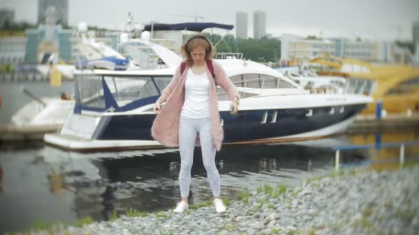 Een meisje met een smartphone op het strand, luisteren naar muziek in hoofdtelefoons, dansen van een jacht en zeilen in de haven. — Stockvideo