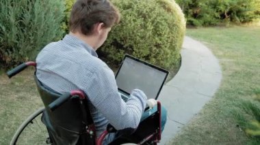 Engelli biri tekerlekli sandalyede oturan ve parkta bir dizüstü bilgisayar üzerinde çalışan