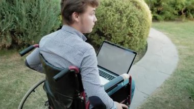 Engelli biri tekerlekli sandalyede oturan ve parkta bir dizüstü bilgisayar üzerinde çalışan