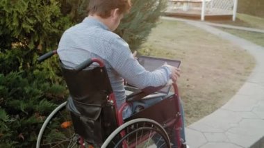 Engelli biri tekerlekli sandalyede oturan ve parkta bir dizüstü bilgisayar üzerinde çalışan