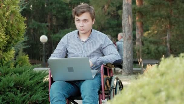 Een gehandicapte man in een rolstoel zit en werken op een laptop in het park — Stockvideo