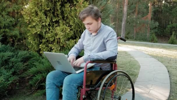 Un disabile è seduto su una sedia a rotelle e lavora su un computer portatile nel parco — Video Stock