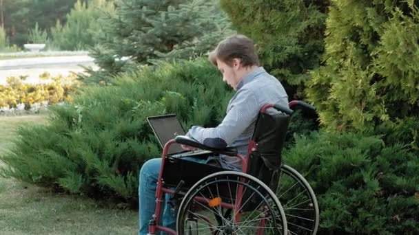 Людина з обмеженими можливостями сидить на інвалідному візку і працює на ноутбуці в парку — стокове відео