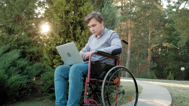 Un hombre discapacitado está sentado en una silla de ruedas y trabajando en un portátil en el parque — Vídeos de Stock