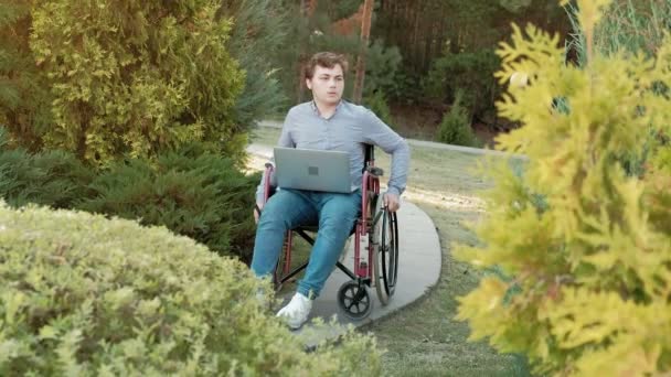 Un handicapé est assis dans un fauteuil roulant et travaille sur un ordinateur portable dans le parc — Video